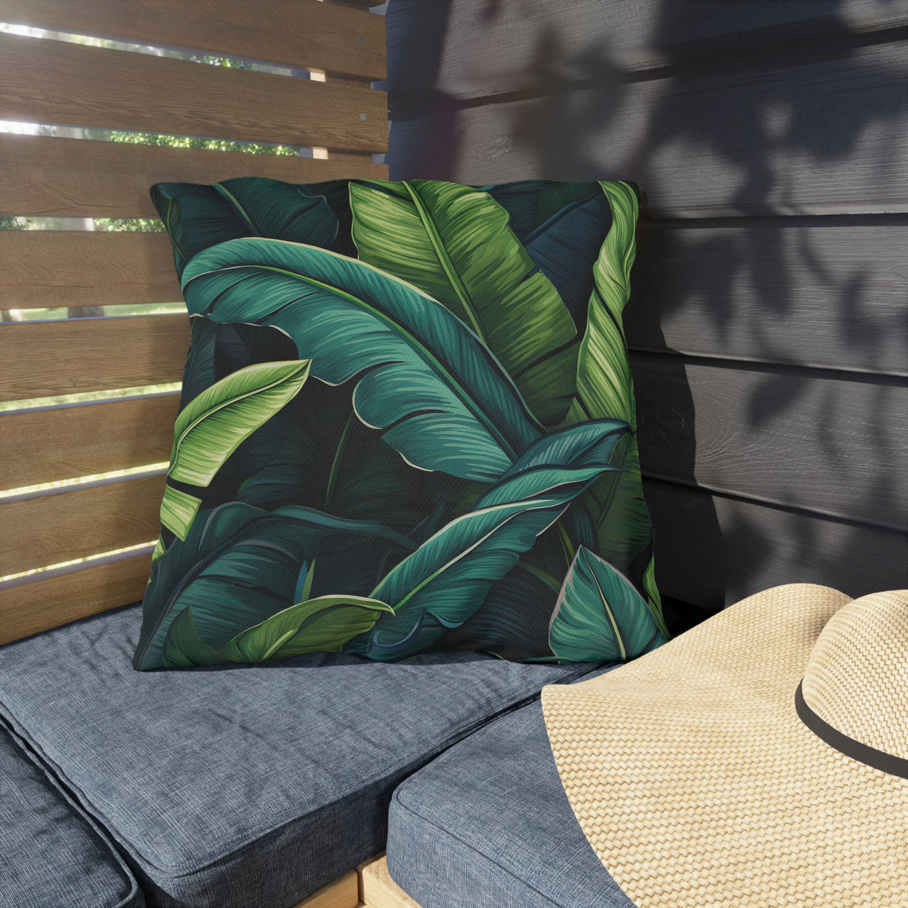 Tropical leaf patterned pillow on a gray cushion with a straw hat on a wooden bench.