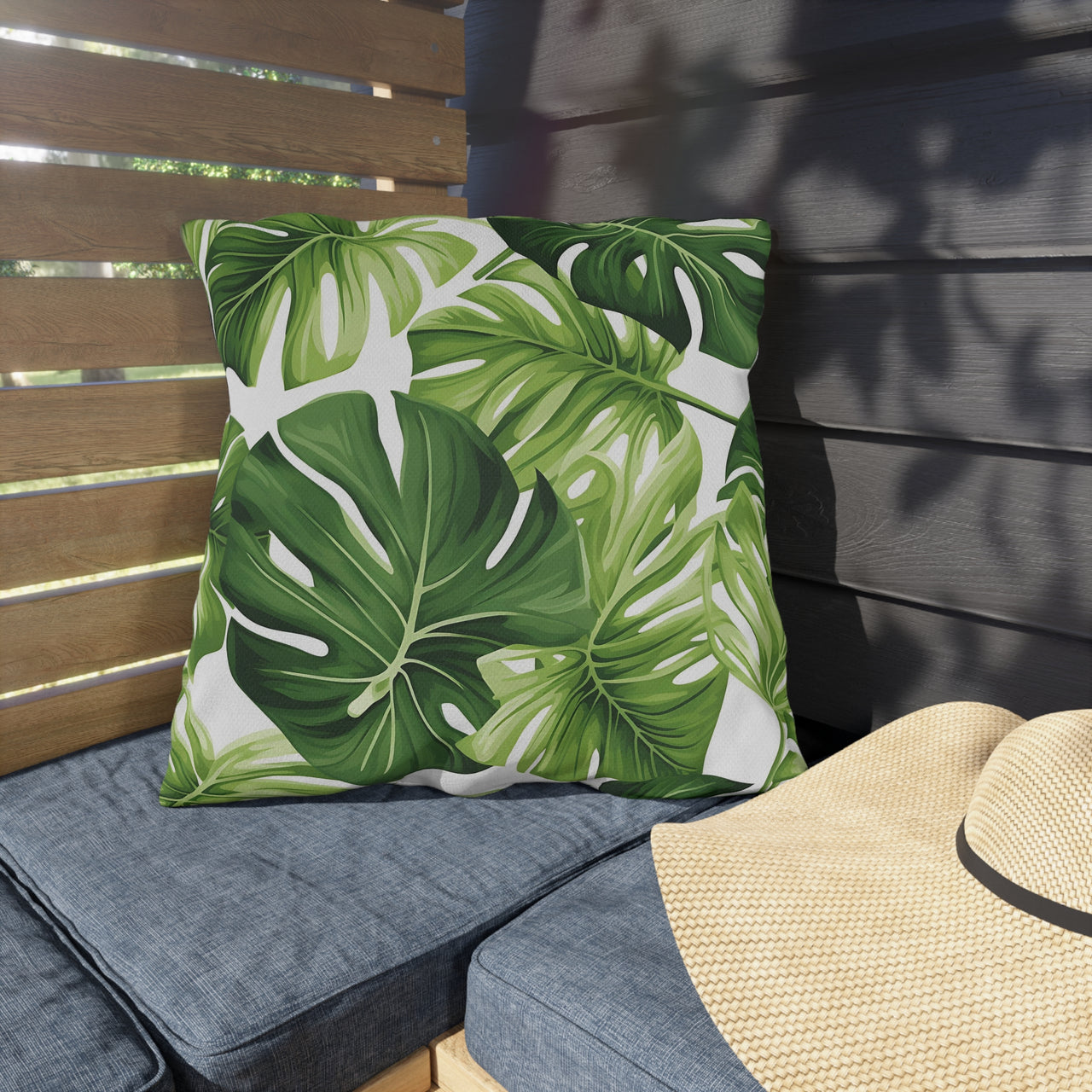 Green leaf-patterned pillow on a gray cushion with a straw hat beside it on a wooden bench.