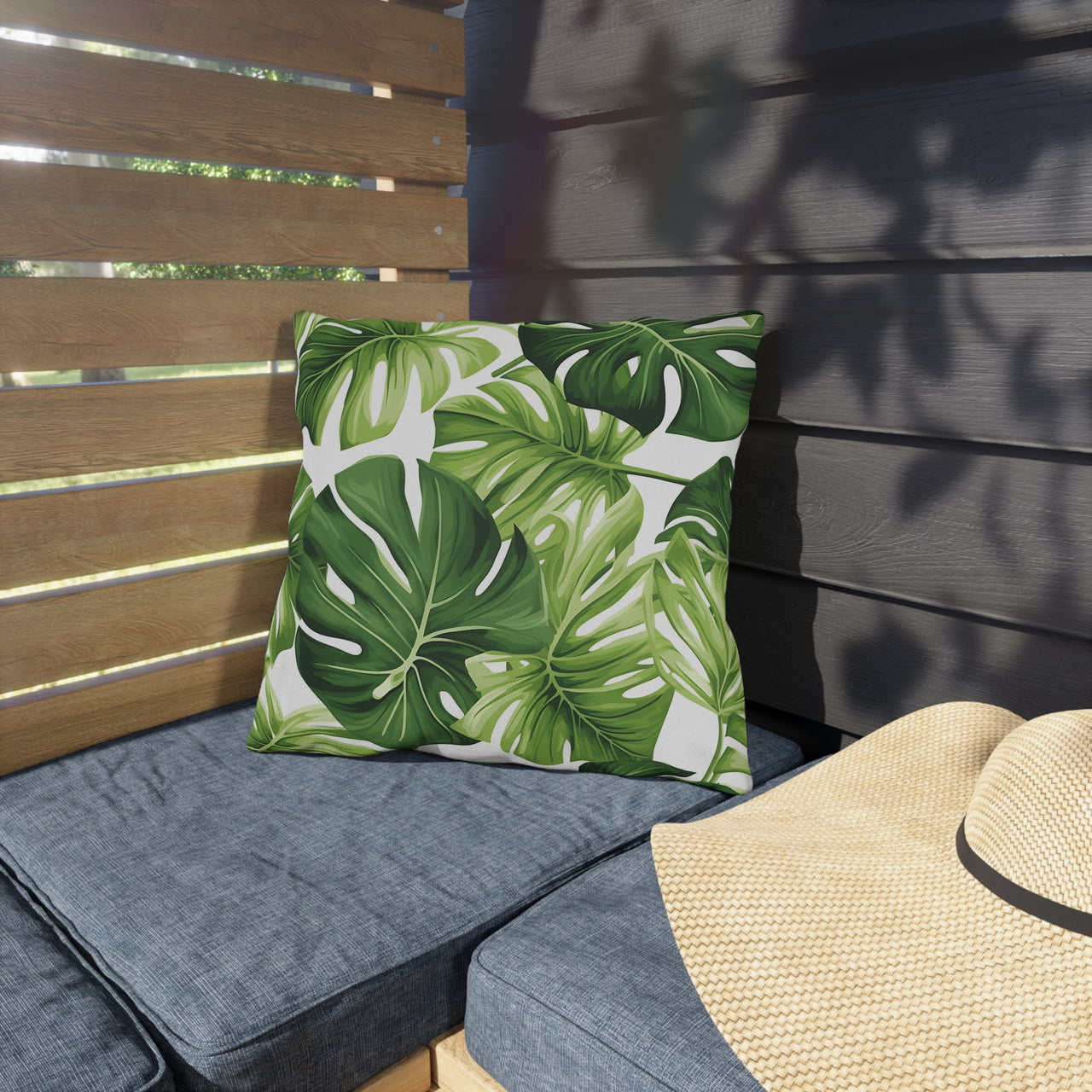 Green leaf-patterned pillow on a gray cushion with a straw hat on a wooden bench.